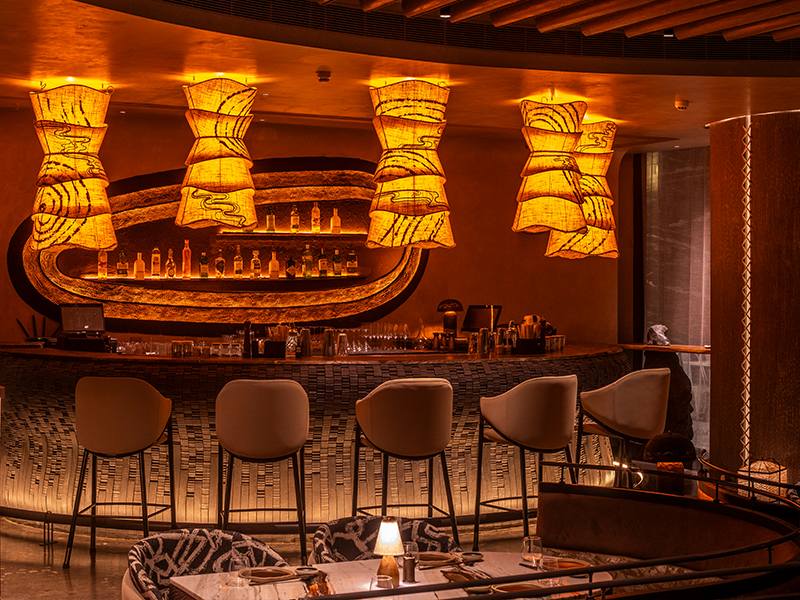 Sculptural details like the resin-carved back bar and fabric-draped pendant lights round out the space, adding an artful finish to the bar’s striking design. ( Image Credit: Donmai)