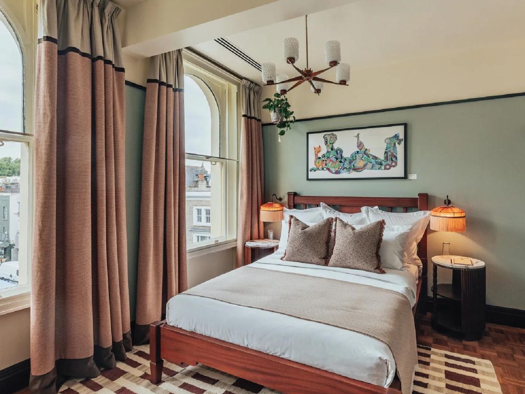 The second bedroom at ‘Permit Room Lodgings’ is a visual delight—anchored by tasselled lamps, Tarini Sethi’s evocative artwork ‘Monday Malaise’ and a window that frames the storied ‘Electric Cinema’ of Notting Hill. Image Credit: (Permit Room Lodgings)