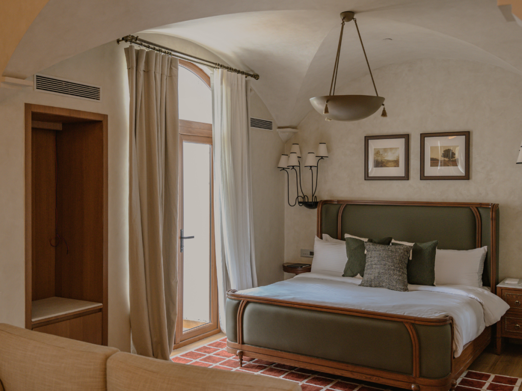 A light-filled guest room styled in warm midcentury modern tones by Humming Tree of Calicut. Clean-lined furniture in rich wood, soft woven rugs, and tactile fabrics create an inviting, layered atmosphere. Artworks — from the eclectic finds of Le Puces in Paris to the intricate craftsmanship of Kala in Kerala — punctuate the walls, adding colour, texture, and a sense of place. (Image Credits: Hotel Irada)
