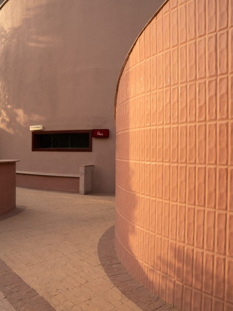 The façade’s sweeping curves catch the late afternoon sun, revealing a medley of textured matte plaster and glossy pink tiles. (Image Credits: Avesh Gaur)